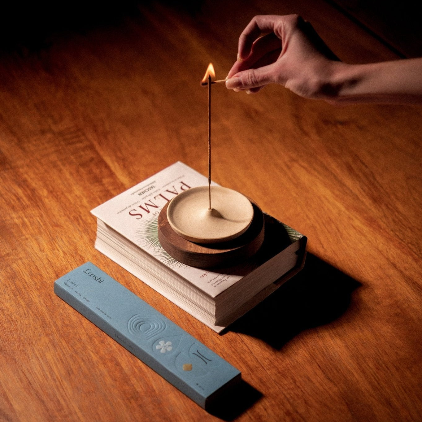 Hand lighting a Looshi Calm incense stick in a round holder placed on stacked books, with the blue product box nearby on a wooden table
