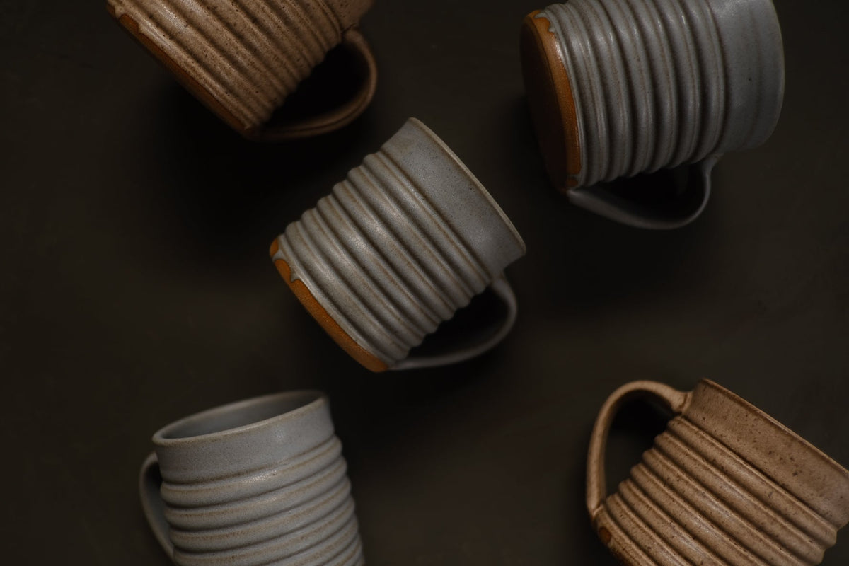 Five Settle Ceramics The Little Mugs in grey and brown arranged at different angles on a dark surface, some partly cropped, showing ribbed ceramic bodies, curved handles and contrasting ochre bases.