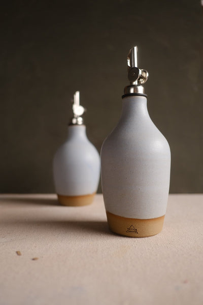 Pale grey Settle Ceramics Wander Oil Bottle in front of a smaller matching bottle on a light tabletop against a dark background, showing softly rounded shoulders, metal pouring spouts and ochre base bands.