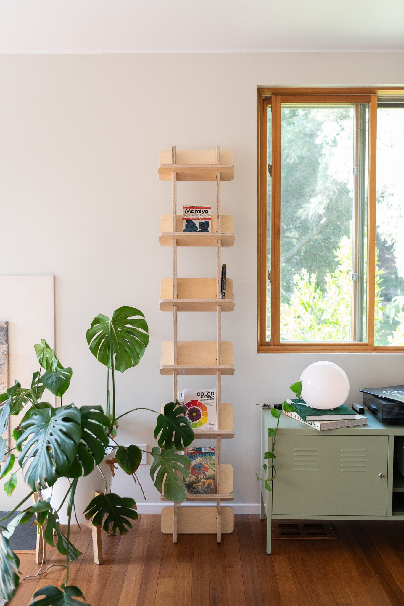 Yankel tall wood bookcase displaying books and small decor beside a green cabinet, round lamp and indoor plants