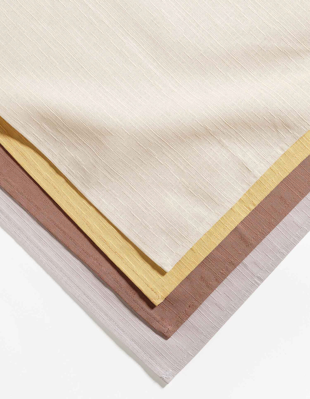 Close-up of stacked Tortuga Forma Dinner Napkins Sabi Mixed Sets in ivory, yellow, dusty rose and pale grey, showing aligned corners, stitched borders and fine linear weave.
