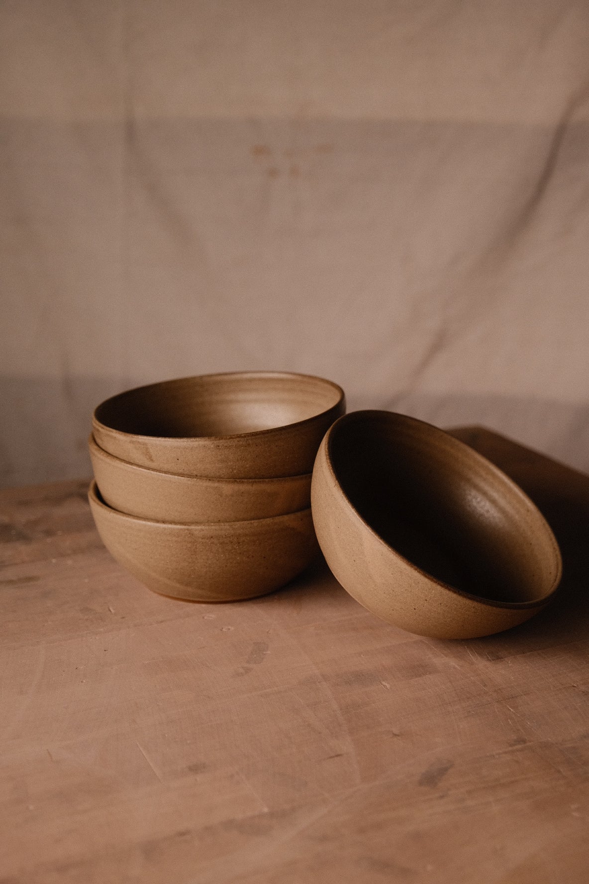 Golden-brown Settle Ceramics Wander Soup Bowls on a wooden tabletop against beige fabric, with three stacked and a fourth leaning beside them, showing rounded ceramic sides, dark rims and subtle concentric lines inside.