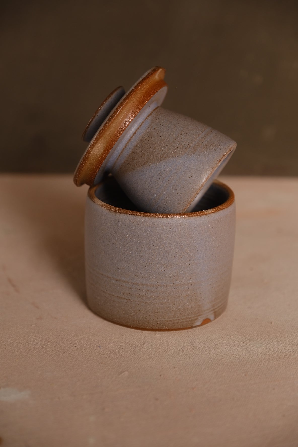 Pale grey Settle Ceramics Butterbell with a subtly speckled cylindrical base and warm brown lid rim, shown with the inner cup tilted outward.