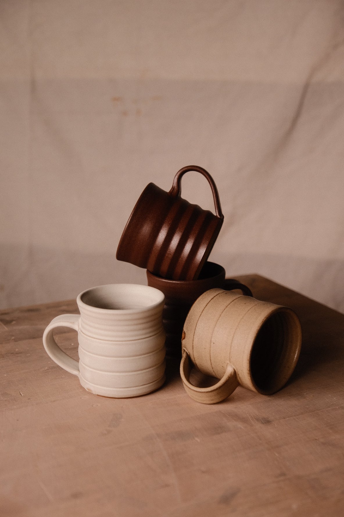 Group of Settle Ceramics The Big Mug on a brown tabletop, with cream and speckled tan mugs in front and dark brown mugs behind, showing cylindrical ceramic bodies, horizontal ridges and curved loop handles.
