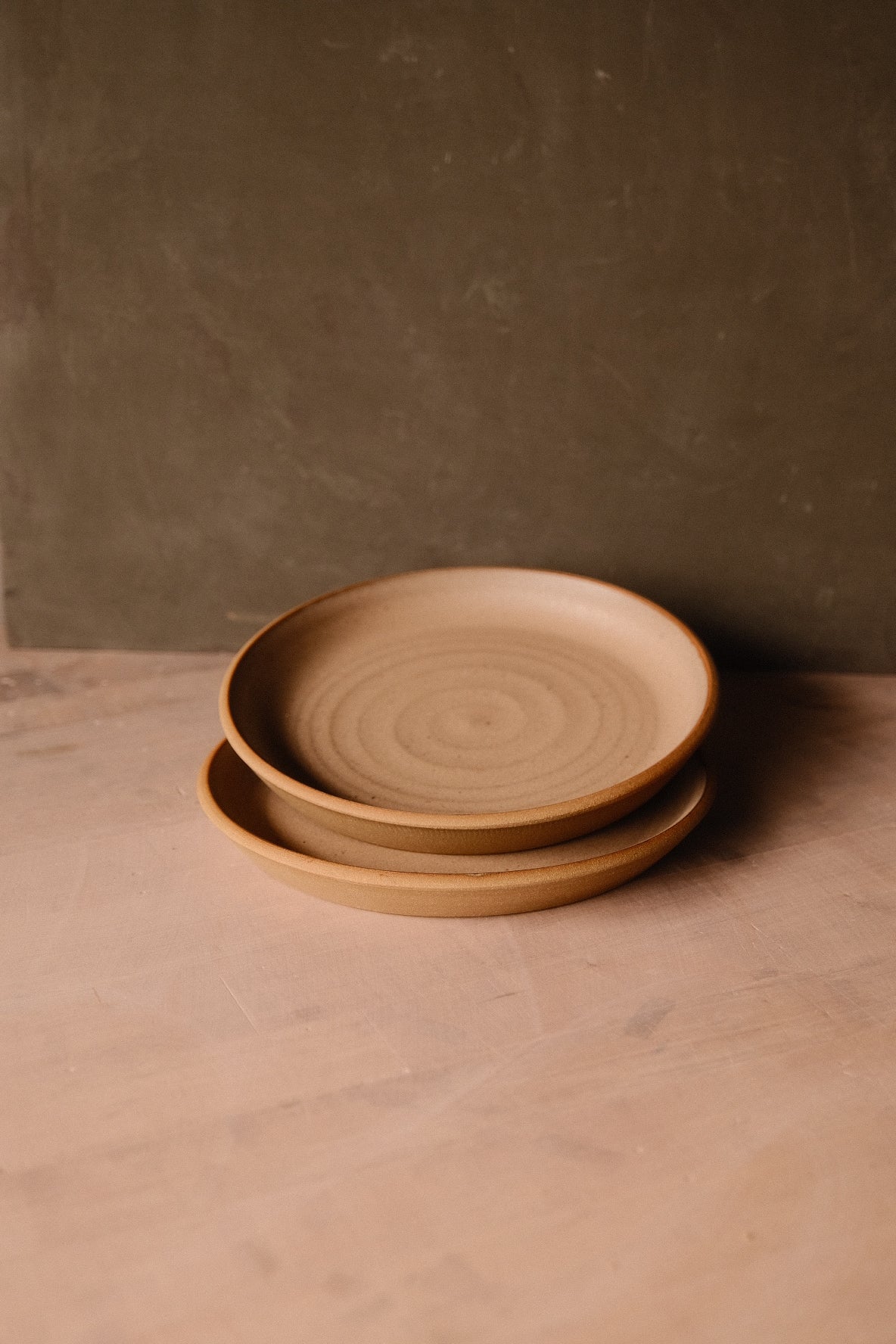 Two warm tan Settle Ceramics Fat Rim Lunch Plates stacked on a brown tabletop, viewed at an angle to show their thick raised rims, shallow centres and concentric surface rings.