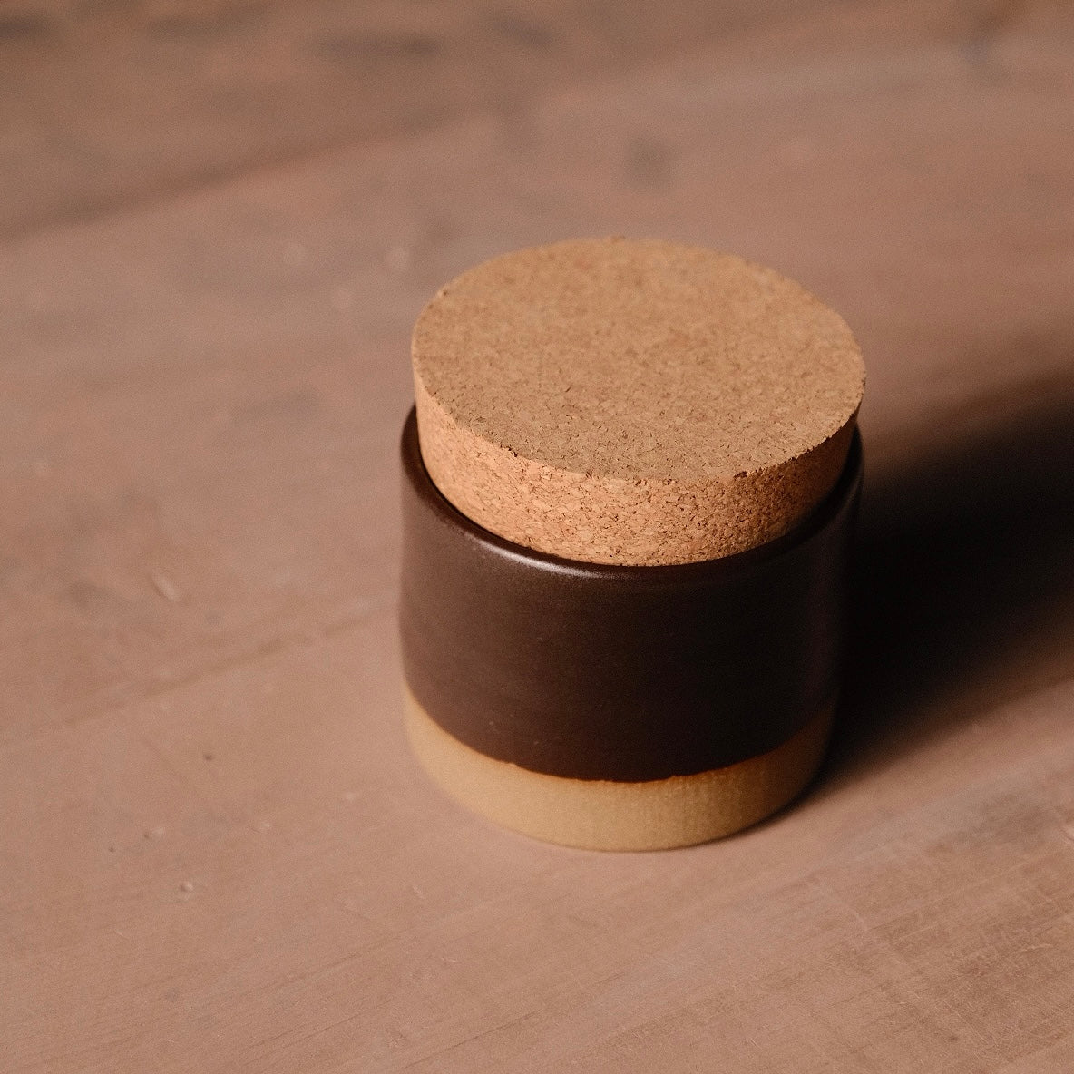 Dark brown Settle Ceramics Wander Salt Jar viewed from above at an angle on a wooden tabletop, showing its short cylindrical ceramic body, contrasting ochre base and thick, flat cork lid.