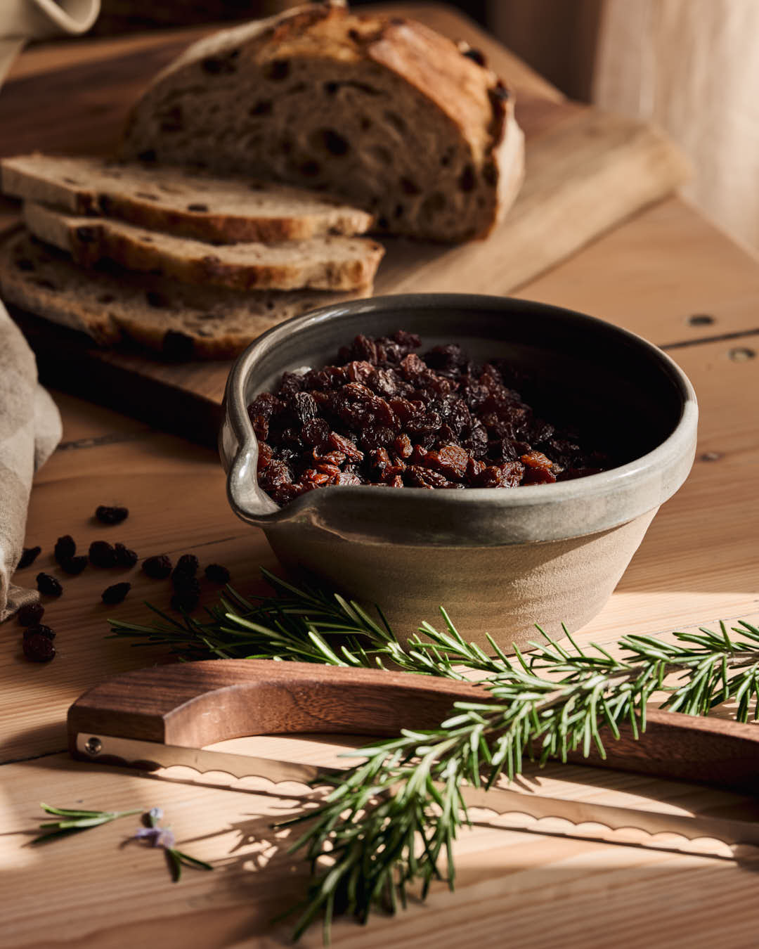 Bread bow resting beside sliced dark bread, a bowl of fruit preserves, chocolate pieces, and fresh rosemary on a wooden surface.