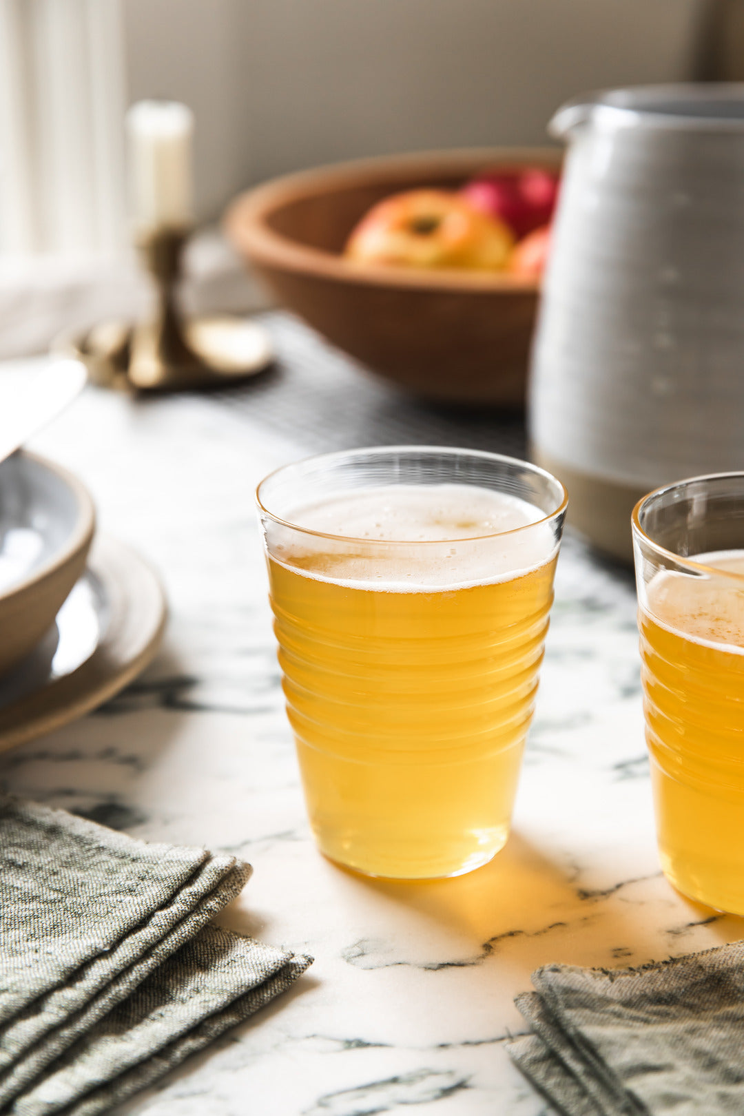 Two clear ridged glasses filled with a golden beverage on a sunlit marble table beside folded green napkins.