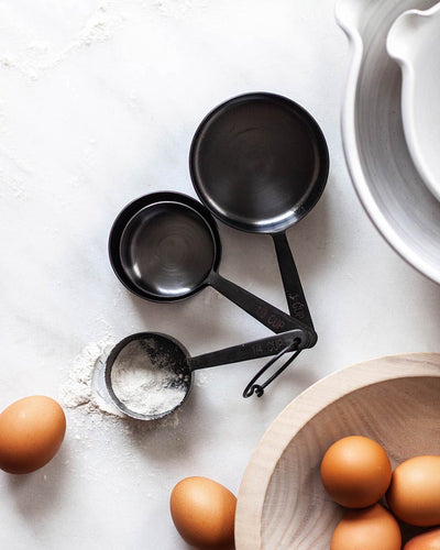 Black measuring cups holding flour beside brown eggs, a wooden bowl, and the curved edge of a white baking dish.