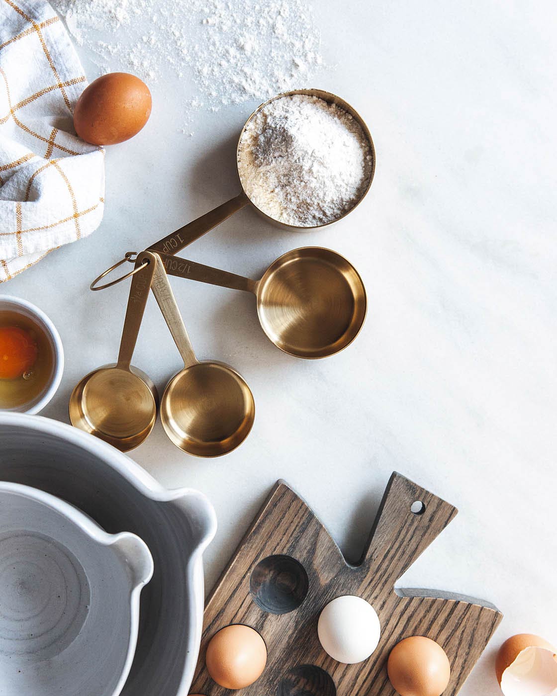 Overhead baking scene with brass measuring cups, flour, eggs, utensils, striped kitchen towels, and ceramic bakeware.