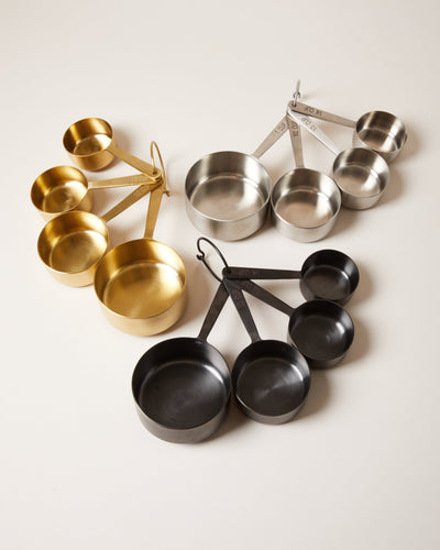 Three nested sets of four round metal measuring cups in brushed brass, stainless steel, and matte black finishes, each joined by a removable ring.