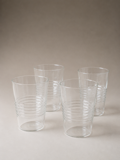Four clear cylindrical drinking glasses with subtly irregular rims and horizontal ridges around their lower halves, arranged on a warm gray surface.