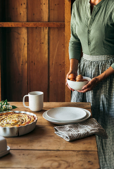 Person holding a bowl of brown eggs beside a rustic table set with a white mug, blue-gray plates, folded linens, and a baked dish.