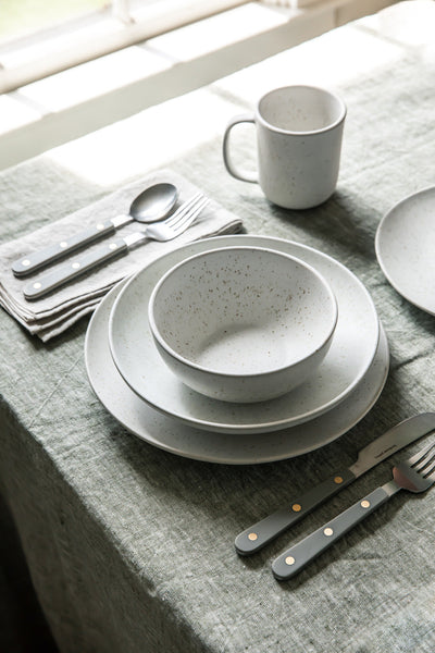 White speckled bowl and stacked plates arranged with a mug, flatware, and gray linen on a sunlit dining table.