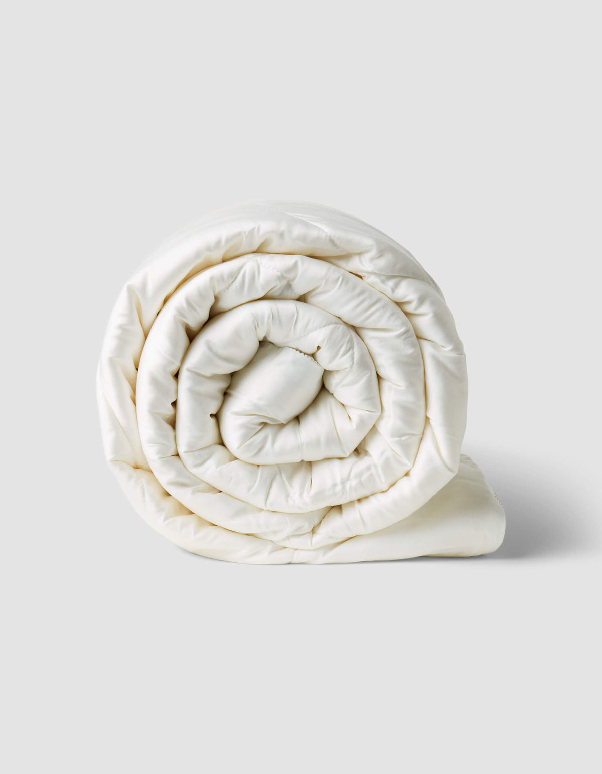 White comforter rolled into a compact spiral on a pale background, showing its padded layers and neatly finished edge.