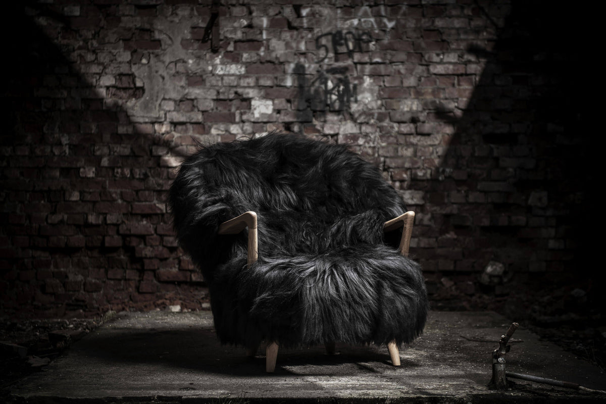 Black Eikund Fluffy Lounge Chair beneath a focused beam of light against a weathered brick wall, with its shaggy silhouette and wood armrests highlighted above a dark, worn floor.