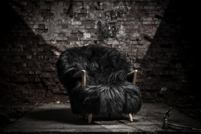 Black Eikund Fluffy Lounge Chair beneath a focused beam of light against a weathered brick wall, with its shaggy silhouette and wood armrests highlighted above a dark, worn floor.