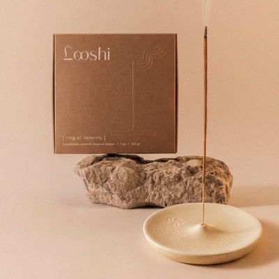 Burning incense stick standing in a cream, shallow bowl-shaped Looshi Ring of Serenity ceramic holder, displayed with natural stone and brown packaging