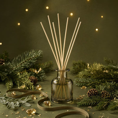 Olive-green Black Cypress diffuser surrounded by evergreen branches, pinecones, leafy sprigs, curled ribbon, and scattered gold accents, with warm lights against a dark green backdrop.