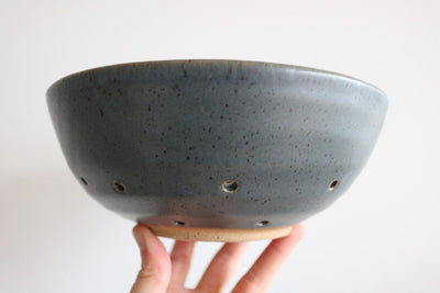Hand holding a dark gray ceramic colander bowl against a white background