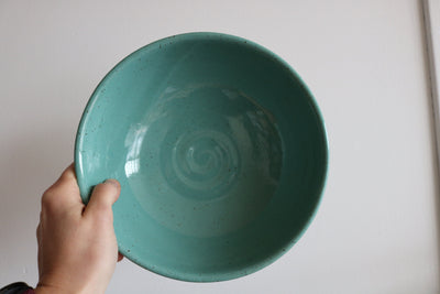 Hand holding a teal ceramic bowl against a plain background by KJ Pottery