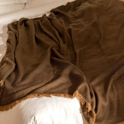Hay Brown merino throw loosely gathered across white bedding, showing its soft fluid drape and fringed border