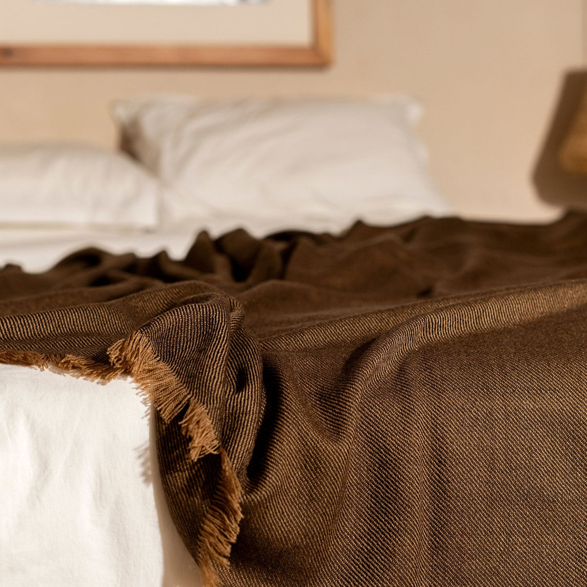 Hay Brown merino throw layered across the foot of a white bed with its textured weave and fringe visible