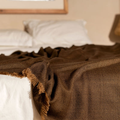 Hay Brown merino throw layered across the foot of a white bed with its textured weave and fringe visible