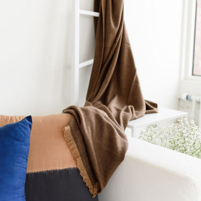 Hay Brown merino throw draped across a white sofa beside tan and navy pillows in a bright room