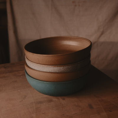 Four Settle Ceramics Wander Serving Bowls stacked on a brown tabletop, with warm brown, pale speckled beige and deep teal glazes, showing wide rounded bodies, upright rims and subtle horizontal ridges.