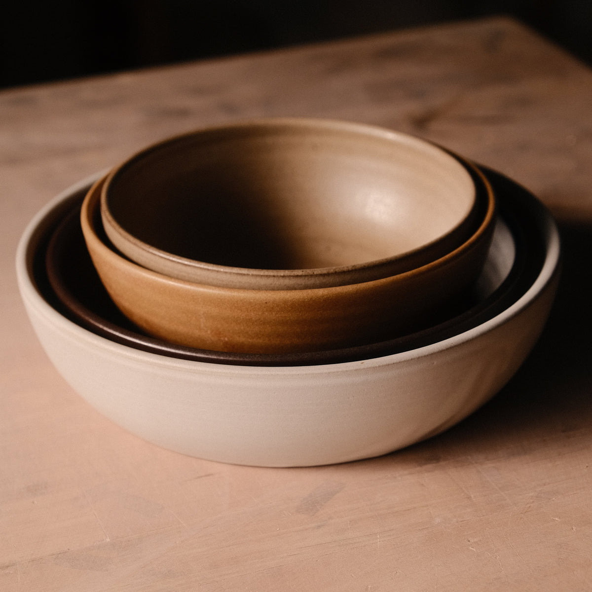 Nested Settle Ceramics Wander Serving Bowls viewed at an angle, with a golden-brown bowl inside darker and pale cream bowls, highlighting their contrasting glazes and rounded ceramic walls.