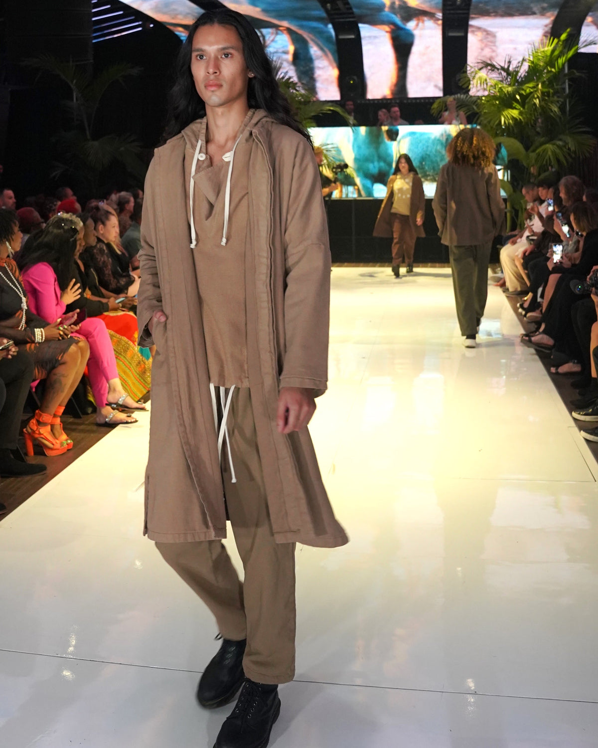 Runway model wearing a tan Passage Jacket over a matching hooded top and loose pants with black shoes.