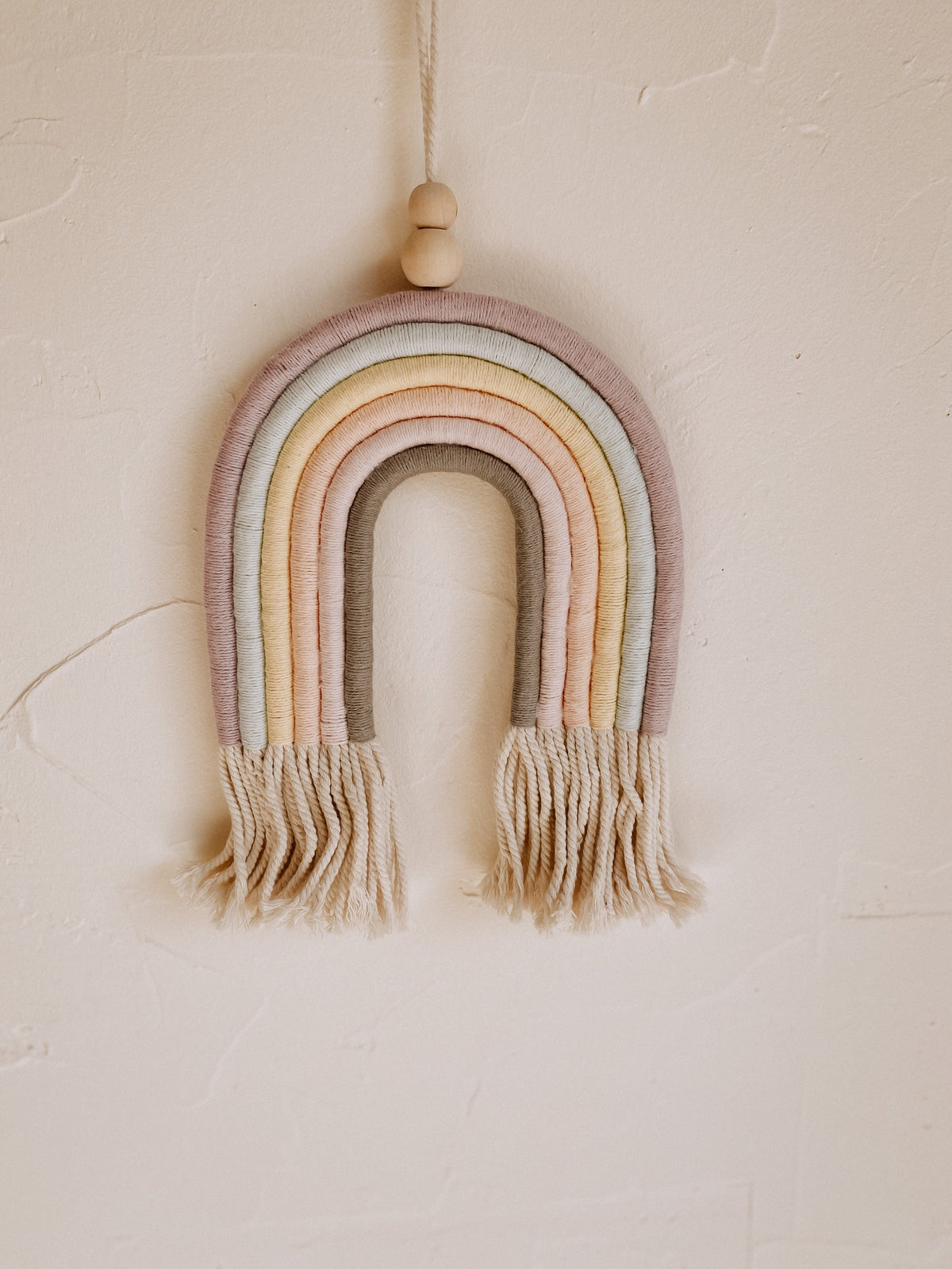 Village Thrive Mia Macrame Rainbow, pastel multicolor cotton, hanging on a wall with a wood bead accent.