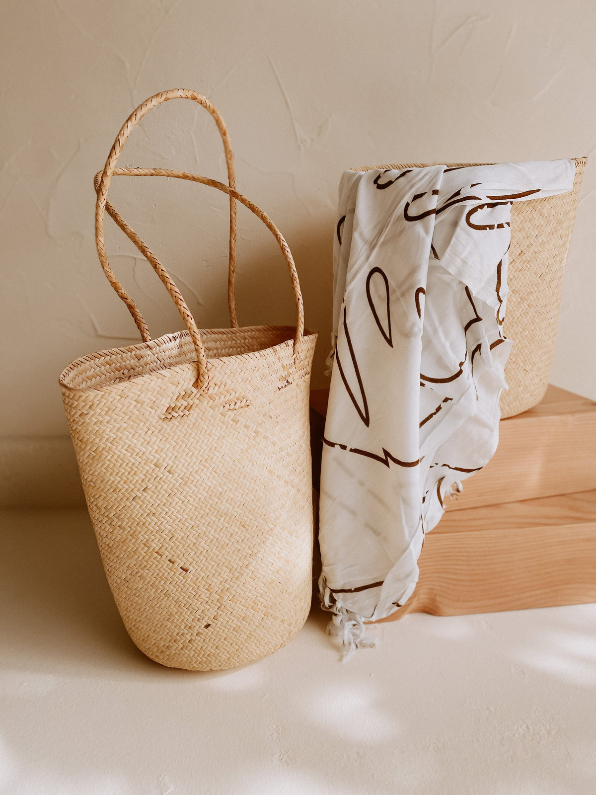 Village Thrive Praya Day Bag, natural rattan, paired with a printed fabric wrap on a wooden bench.