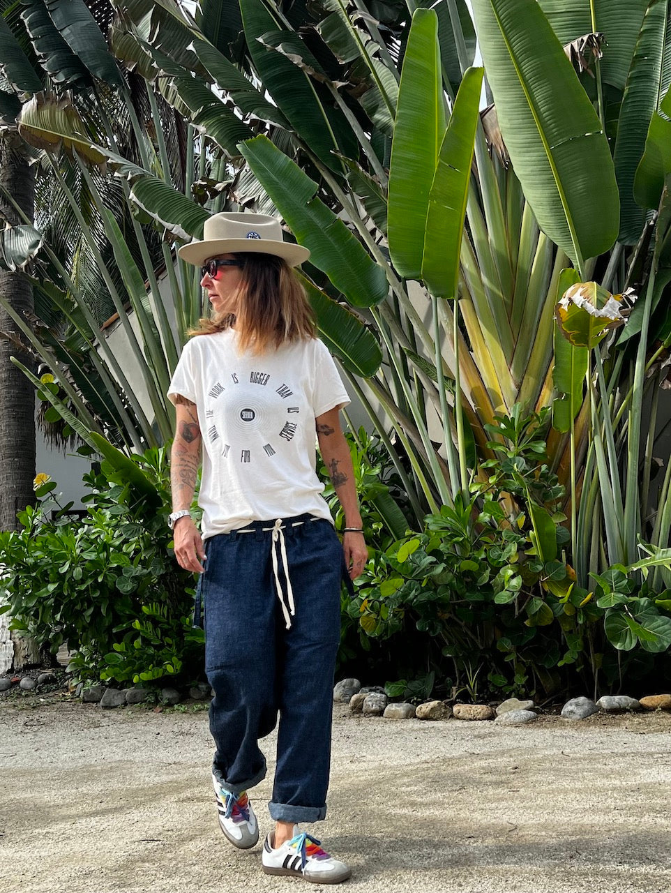 Full-length outdoor view of the model wearing the graphic Service Tee with navy pants and a beige hat among tropical plants.