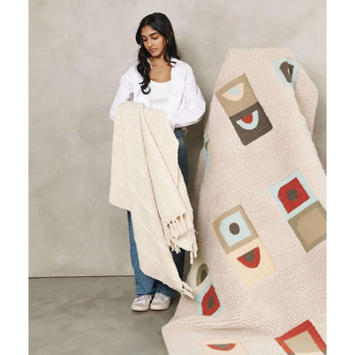 Woman holding the Sea Shell White throw in front of a multicolored geometric rug to show its neutral tone and fringed border