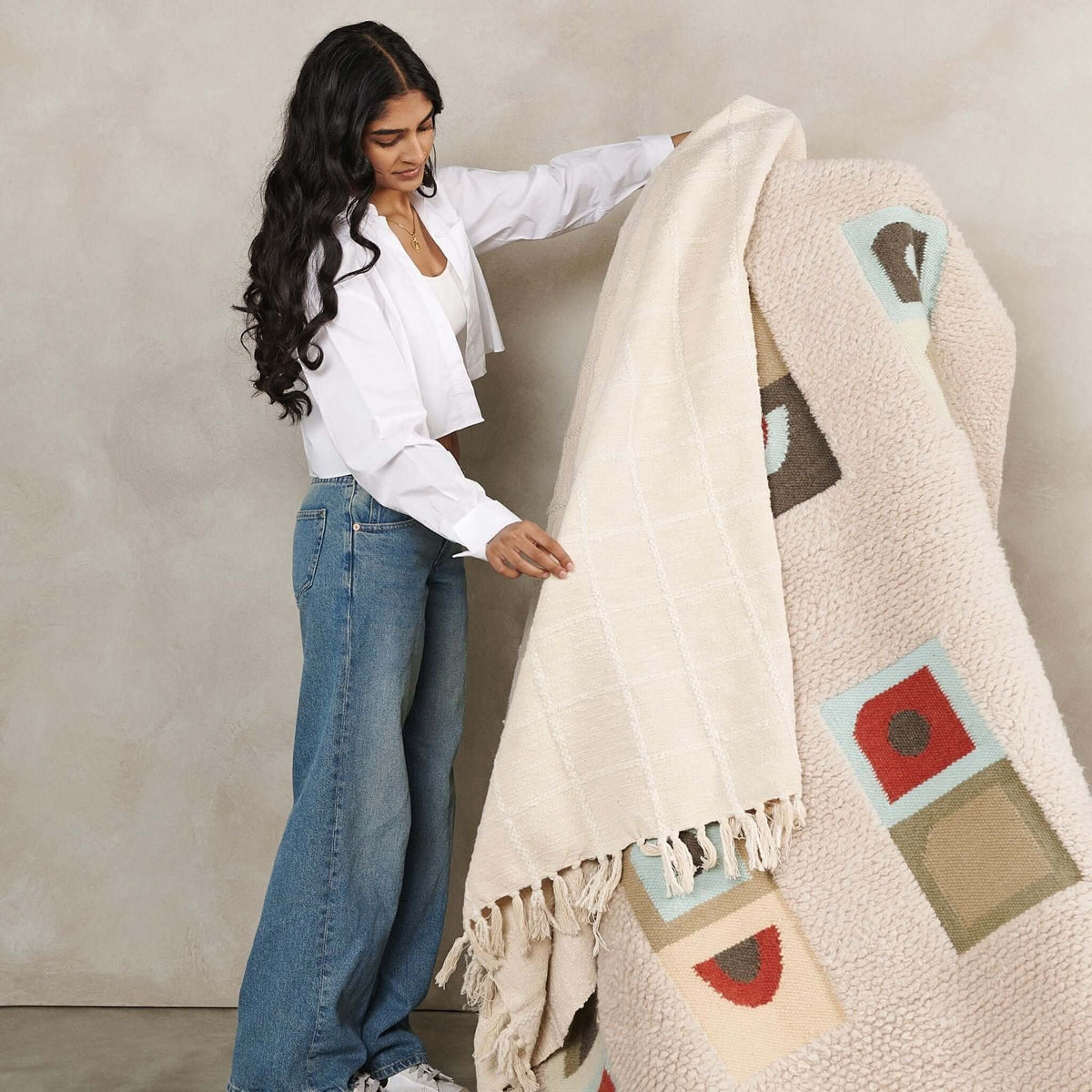 Woman unfolding the Sea Shell White handloom throw beside a hanging rug with colorful geometric motifs