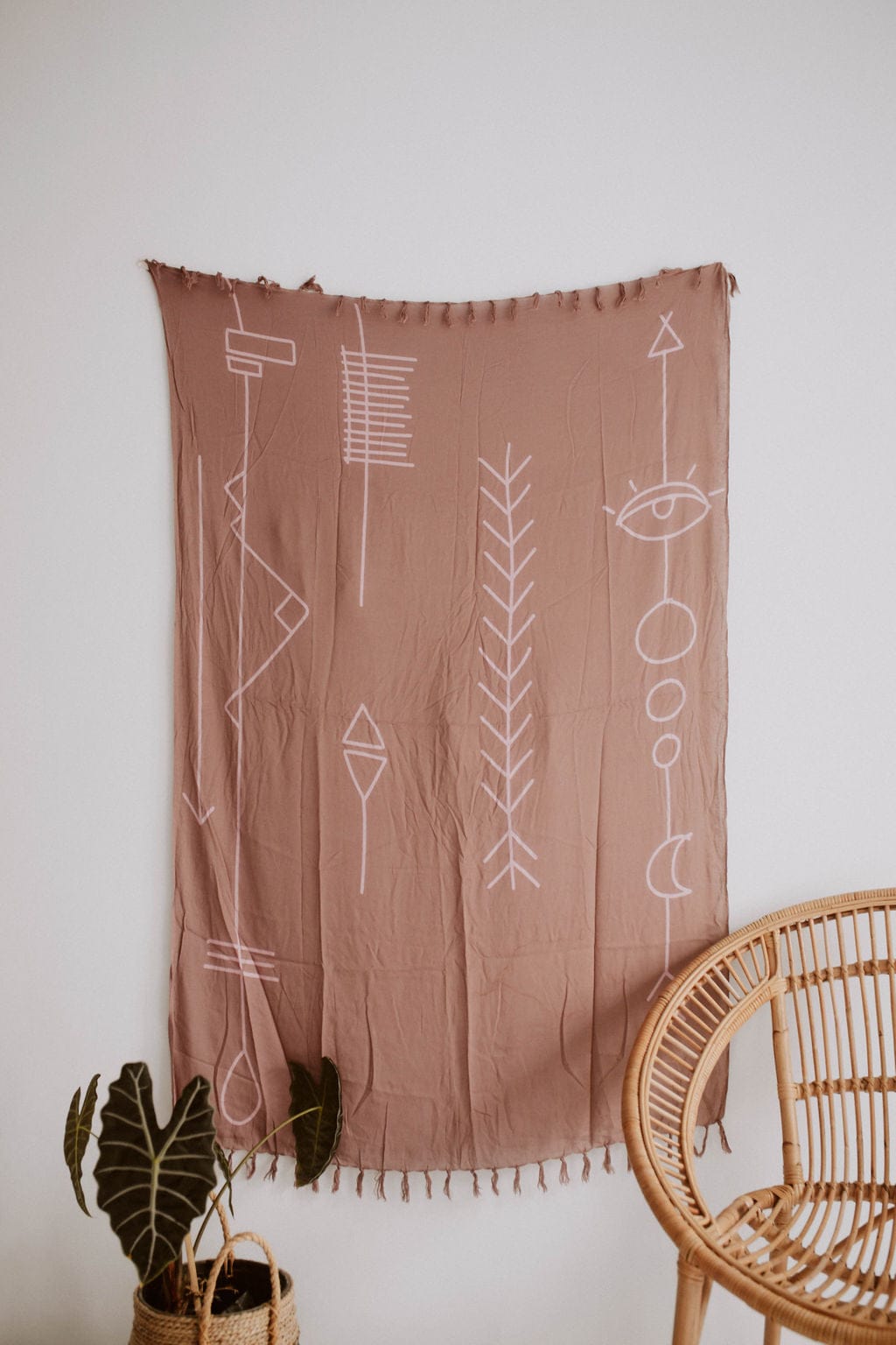 Village Thrive Abstract Tapestry, dusty rose rayon, hanging above a rattan chair with a potted plant.