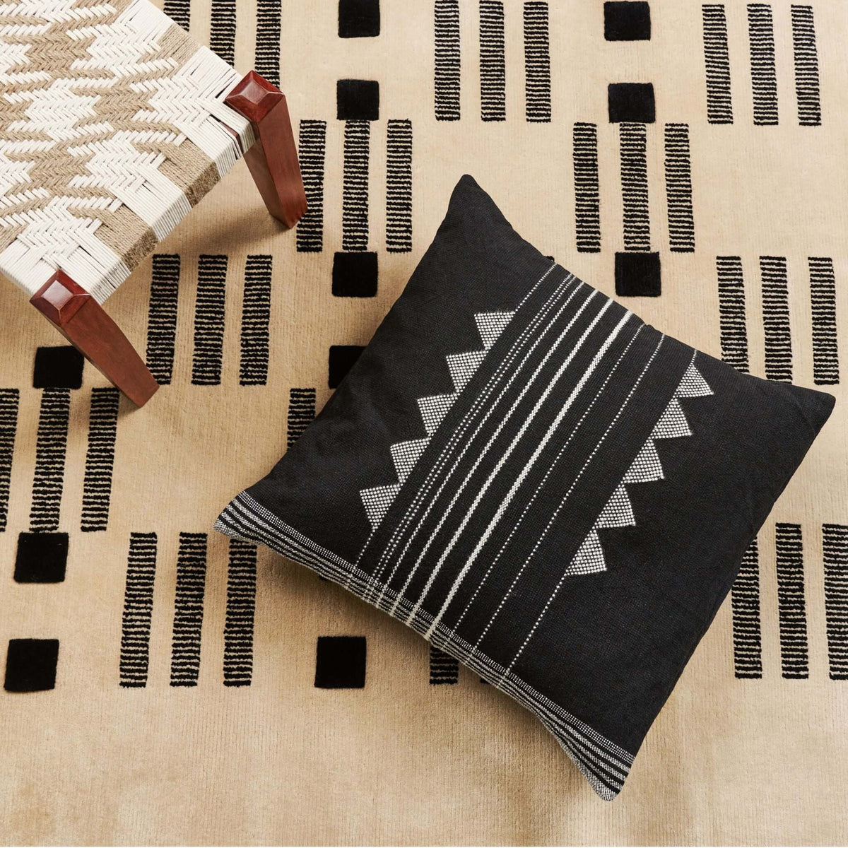 Kora Black artisanal pillow displayed on a cream rug with black geometric markings and layered woven textiles