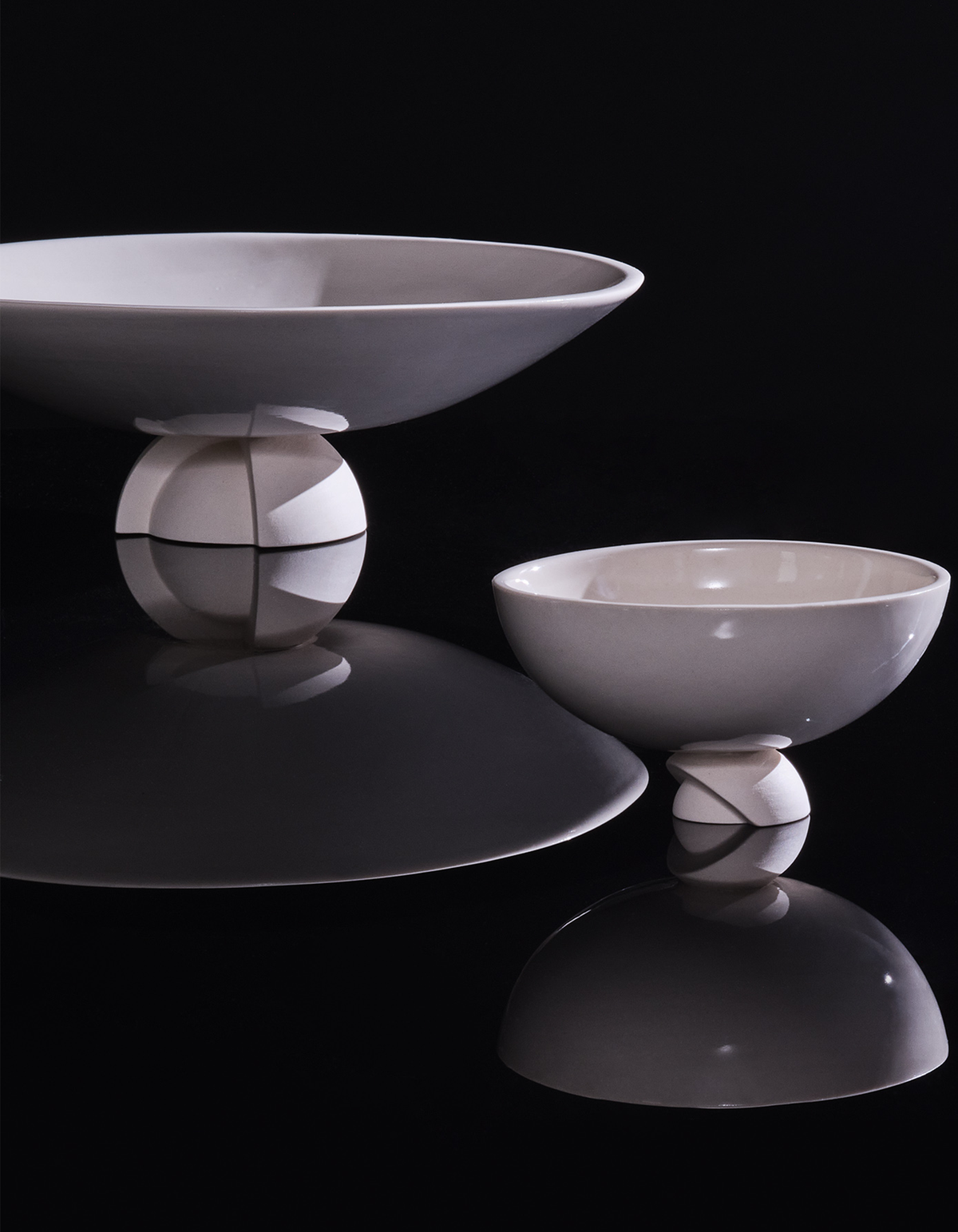 White Tortuga Forma Kaolin Rice Bowl beside a larger shallow bowl on a glossy black surface, with reflections highlighting their curved profiles and sculptural segmented feet.