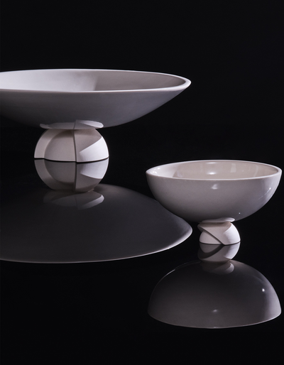 White Tortuga Forma Kaolin Rice Bowl beside a larger shallow bowl on a glossy black surface, with reflections highlighting their curved profiles and sculptural segmented feet.