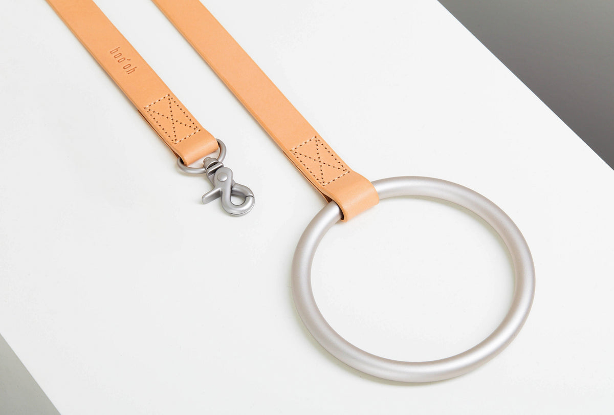 Close-up of the peach leather-like Lumi Leash with a brushed-silver swivel clasp, perforated stitched details, and oversized circular metal handle.