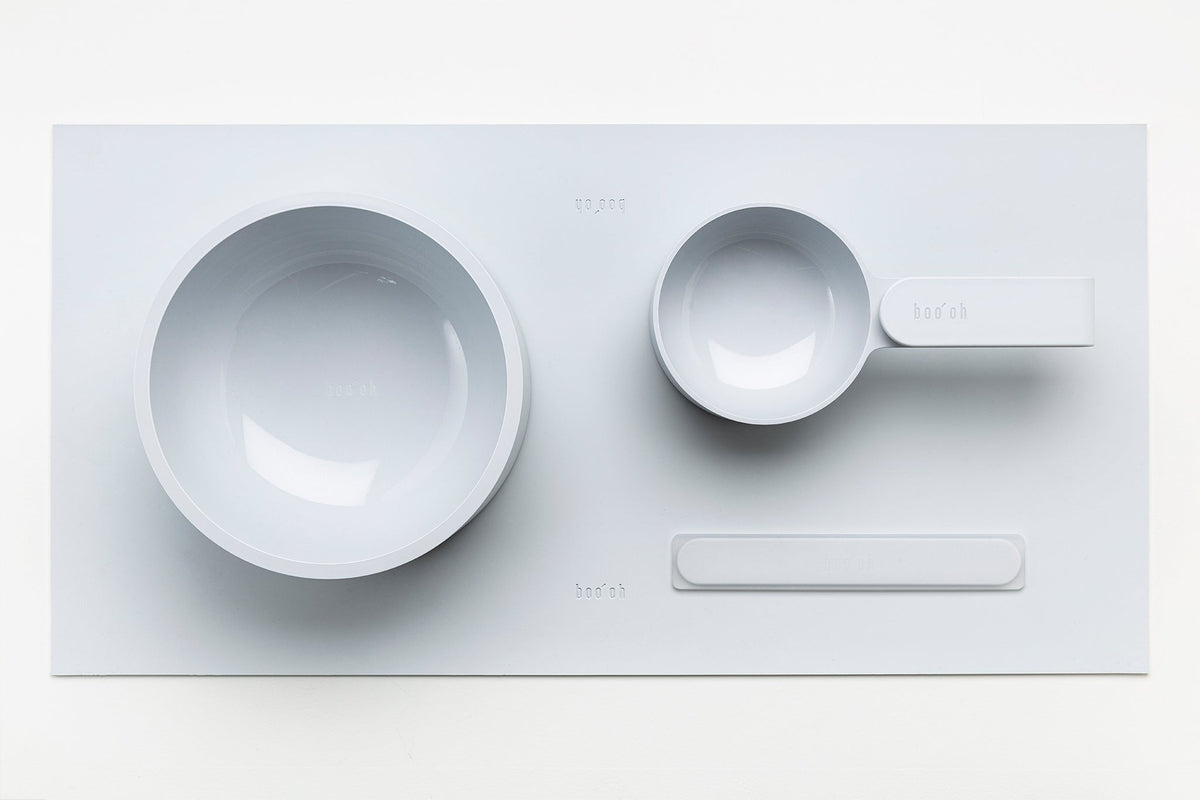 Coordinated pale gray pet-feeding set with two round Mogo Bowls, a spoon, and the slim white spatula arranged on a rectangular mat.