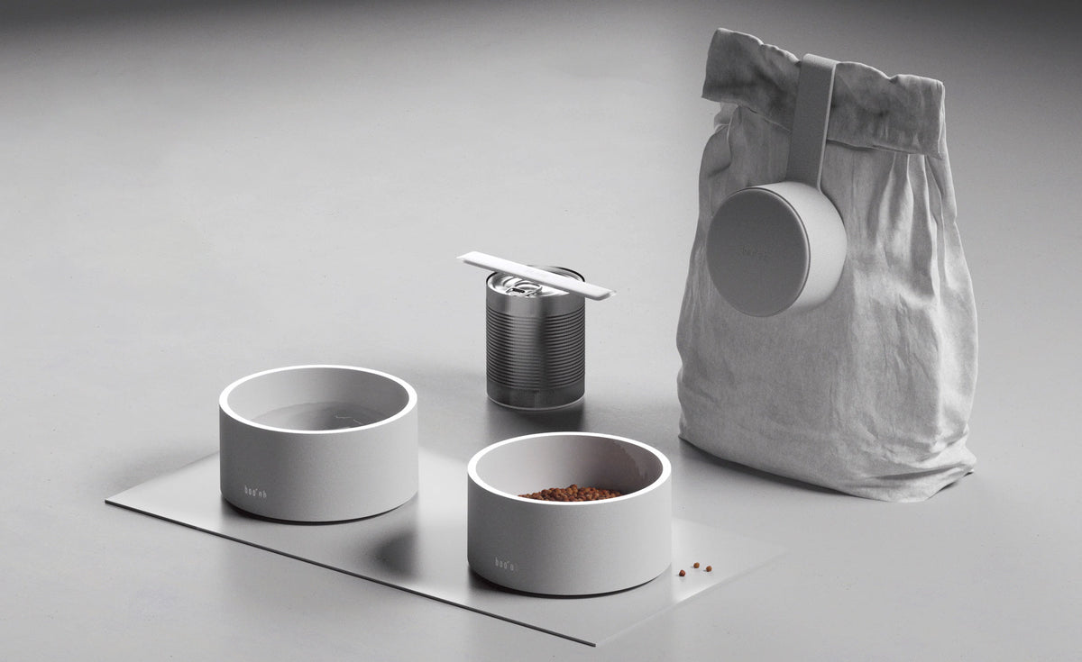 Coordinated pet-feeding setup with two Mogo Bowls, a textured treat container, and a white pet-food bag on a rectangular mat.