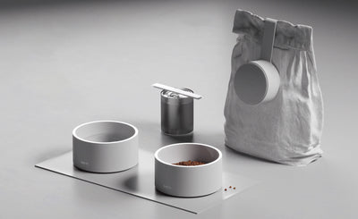 Coordinated pet-feeding setup with two Mogo Bowls, a textured treat container, and a white pet-food bag on a rectangular mat.