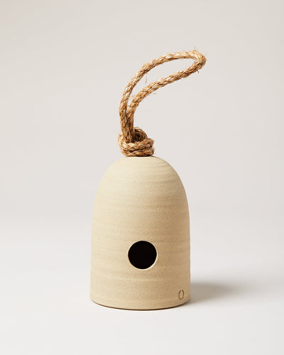 Bell-shaped cream stoneware bird shelter with a round entrance hole, subtle horizontal pottery lines, and a thick natural rope loop knotted through the top.
