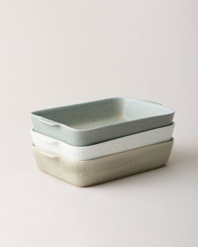 Three rectangular stoneware bakers stacked in soft blue-gray, speckled white, and warm stone glazes, each with rounded corners, gently flared sides, and small integrated side handles.