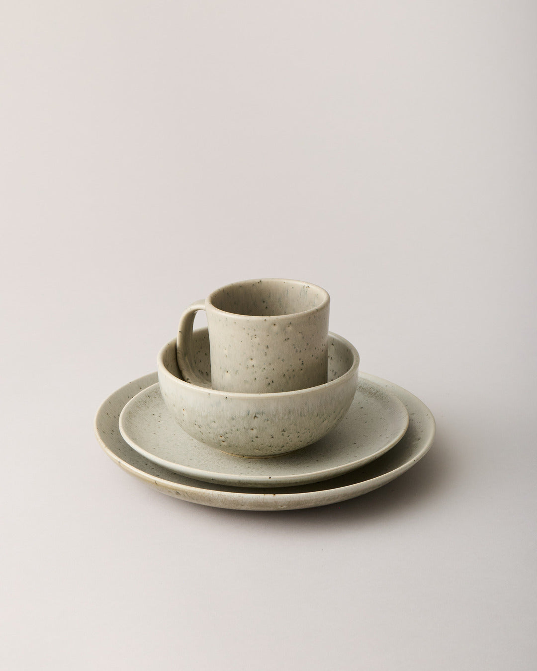 Warm oatmeal speckled mug and bowl stacked on two coordinating plates against a neutral studio background.