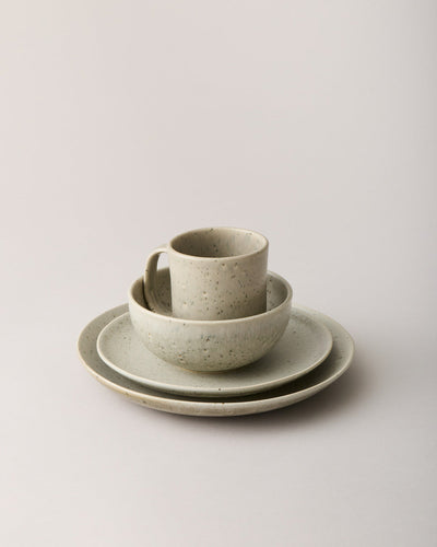 Warm oatmeal speckled mug and bowl stacked on two coordinating plates against a neutral studio background.
