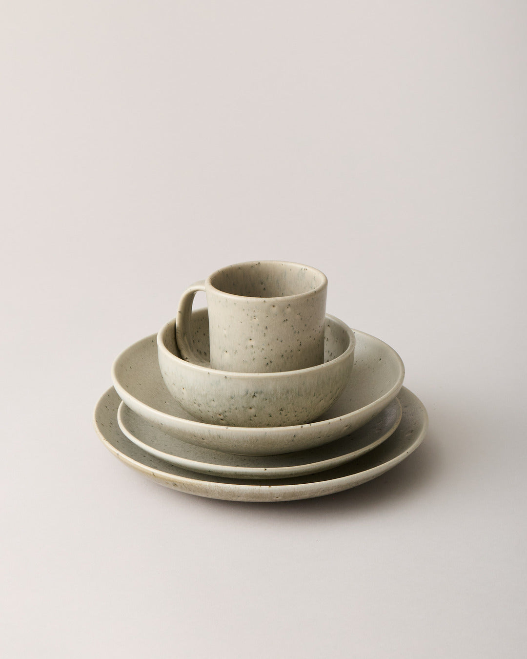 Five-piece stoneware place setting in a warm oatmeal glaze with dark speckles, featuring a straight-sided mug nested inside a bowl above three stacked plates.